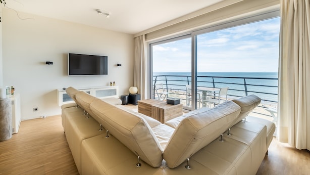 A modern living room with a large sofa, a flat-screen TV, and a balcony with a view of the sea. - Apartment by Zeedijk With Sea View (Koksijde)