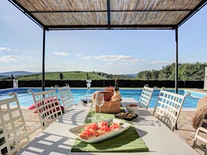 An outdoor dining area with a pool, white chairs, a table with food and drinks, and a view of the landscape. - Villa With a Private Swimming Pool and Tennis Court (Gaiole in Chianti)