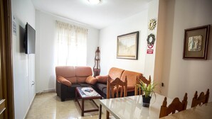 A living room with a sofa, a coffee table, a dining table with chairs, a television mounted on the wall, and a window with curtains.