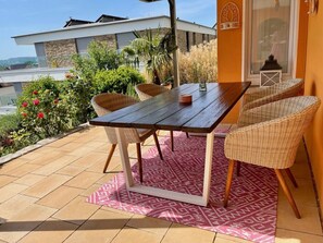 A patio with a wooden table and wicker chairs, a patterned rug, and a view of a building in the background.