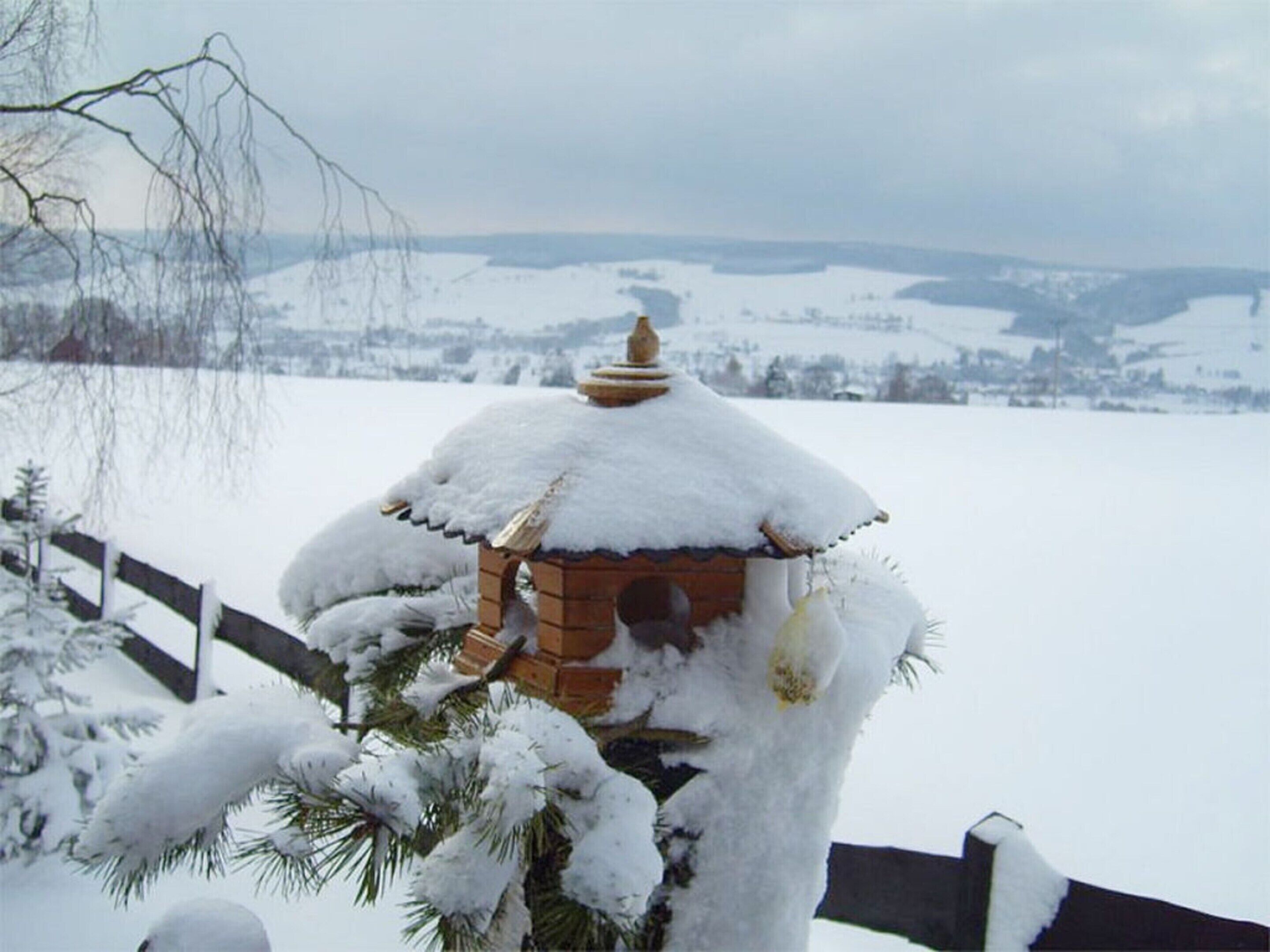Een met sneeuw bedekte gazebo met een fundering van baksteen en een rieten dak, omringd door sneeuwzware bomen en een houten hek.