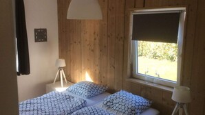 A bedroom with a wooden wall, a window with a blind, a bed with patterned bedding, a bedside lamp, and a picture frame on the wall. - Holiday Home De Zilvermeeuw (Julianadorp)