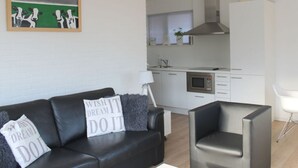 A modern living room with a black leather sofa, a grey armchair, and a painting of cows on a green field. - Holiday Home De Zilvermeeuw (Julianadorp)
