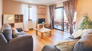 A cozy living room with a sofa, a coffee table, and a TV. There's a balcony with a table and chairs, and a plant on the right. - Holiday Apartment DEA Vista (Medulin)