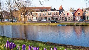 A calm lake with purple crocuses in the foreground, surrounded by traditional European-style buildings with red roofs and a row of parked cars along the shore. - Holiday Home in Alkmaar by Canal (Alkmaar)