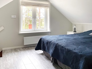 A bedroom with a sloped ceiling, a large window, a bed with a blue quilt, a bedside table with a lamp, and a radiator. - 4 Star Holiday Home in Bro (Bro)