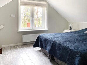 A bedroom with a sloped ceiling, a window with a view of trees, a bed with a blue quilt, a bedside table with a lamp, and a radiator. - 4 Star Holiday Home in Bro (Bro)