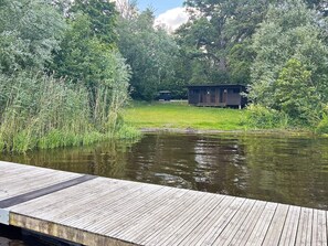 Une jetée en bois s’étendant sur une étendue d’eau calme, entourée d’une végétation luxuriante et surmontée, au loin, d’une petite construction en bois.