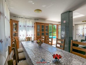 A dining area with a long table, chairs, a cabinet with glass doors, and a view of a swimming pool. - Adalene Comfortable Holiday Residence (Liznjan)