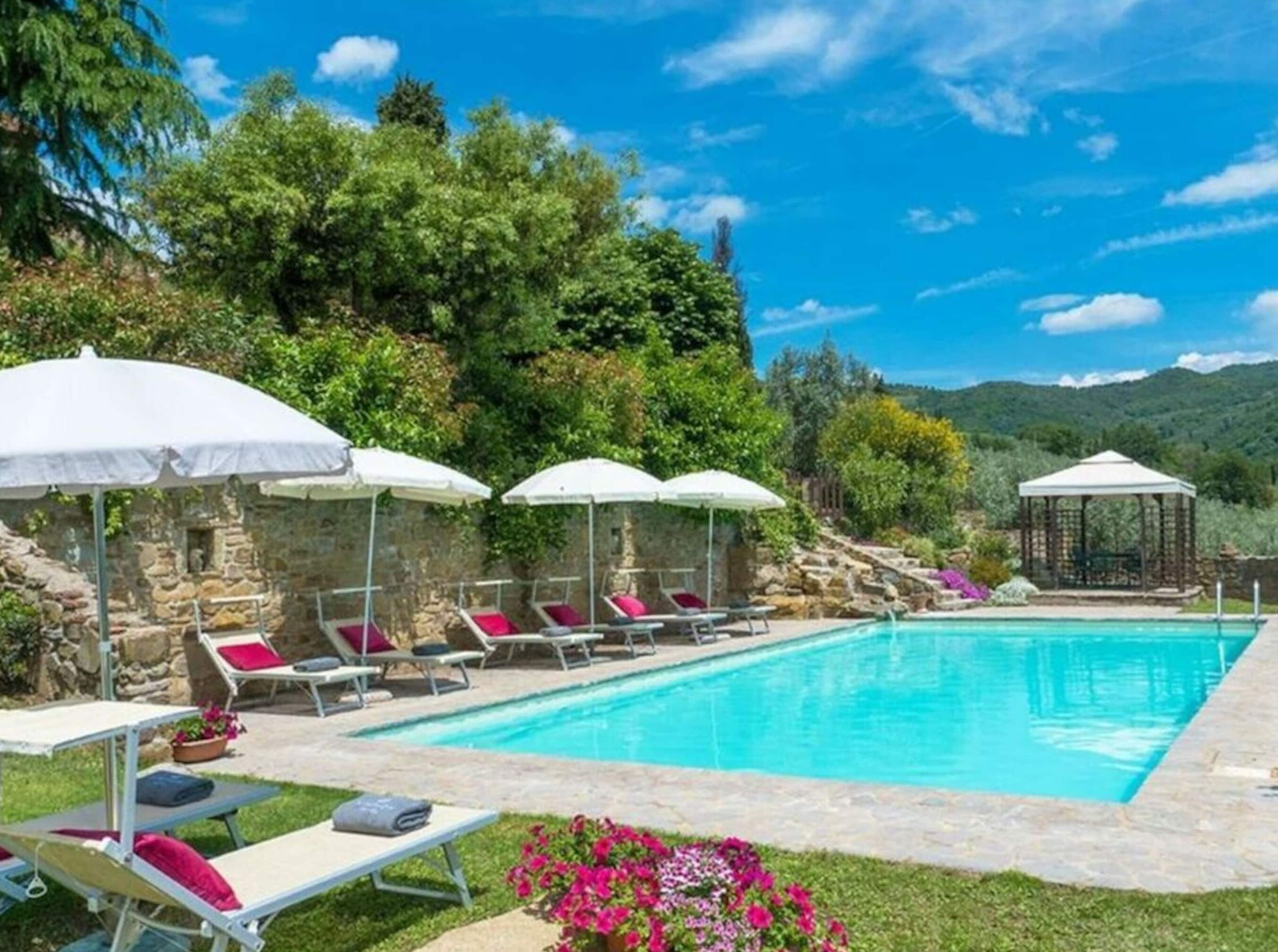 A swimming pool with lounge chairs and umbrellas, surrounded by greenery and stone walls.