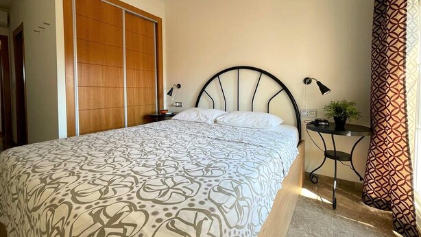 Un dormitorio con cama, mesita de noche, lámpara, planta en maceta y puerta corredera.