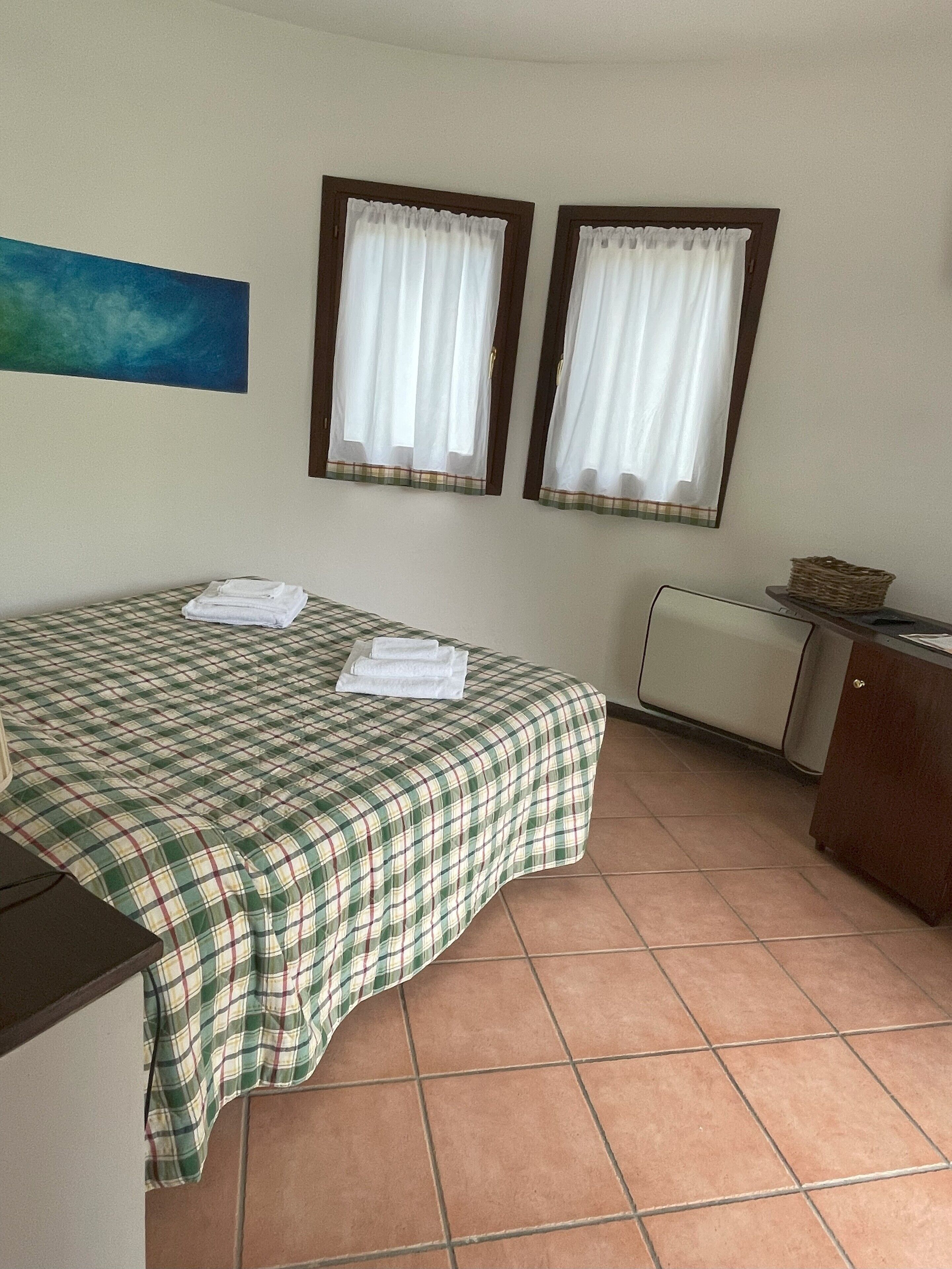 A room with a bed, two windows with curtains, a radiator, and a painting on the wall.