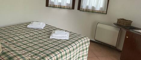 A room with a bed, two windows with curtains, a radiator, and a painting on the wall.