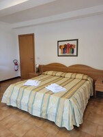 Habitación de hotel con cama, cabecera de madera, mesita de noche, cuadro con flores y extintor.