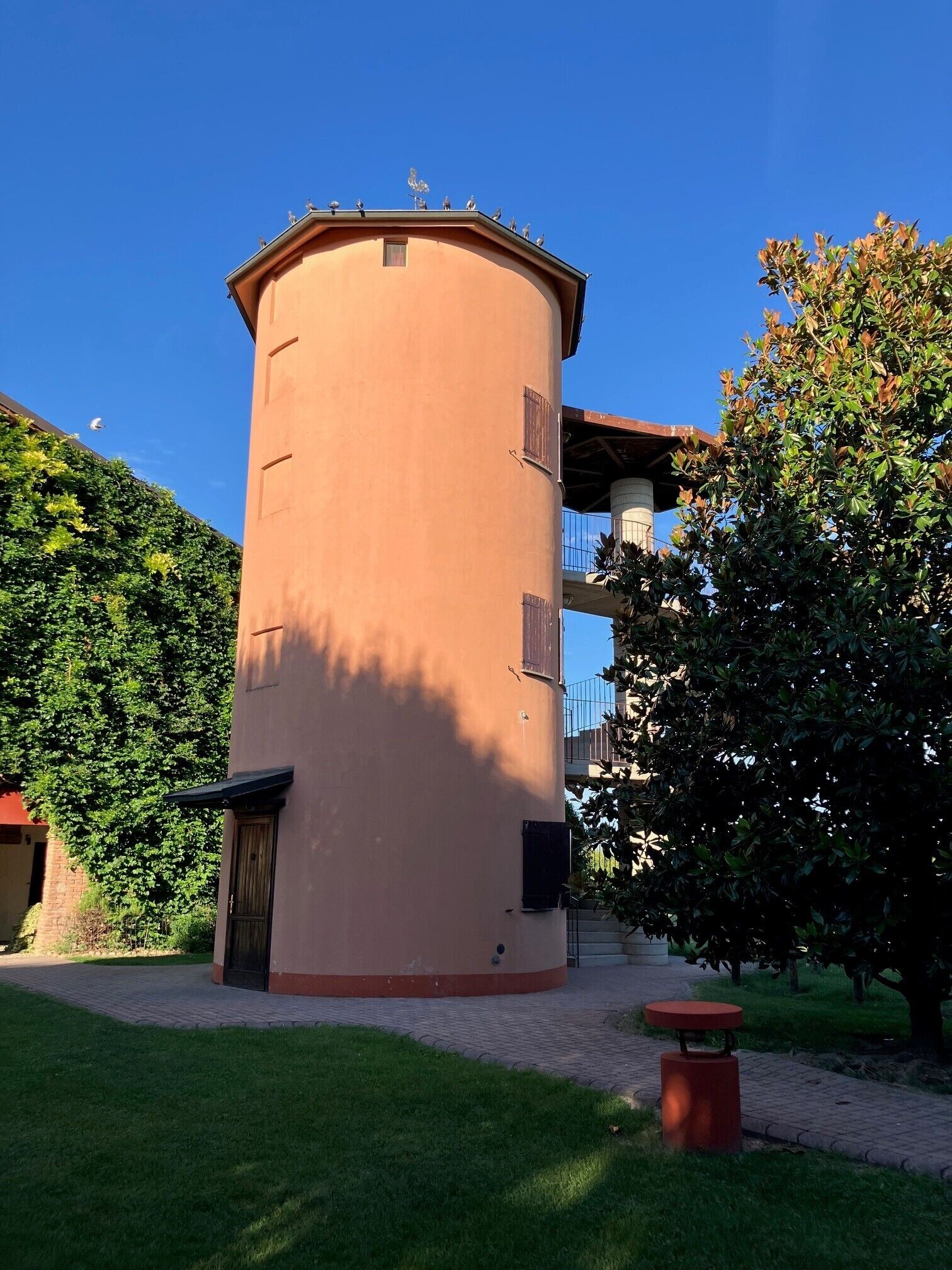 A cylindrical building with a flat roof, surrounded by greenery and a paved walkway.