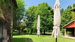 Un jardin avec un chemin de pierres, de l’herbe verte et des arbres. On y trouve deux parasols et un bâtiment avec un toit rouge.
