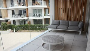 A modern balcony with a grey sofa, a round wooden table, and a view of a multi-story building with outdoor seating. - Apartment in Nieuwpoort Near the Marina (Nieuwpoort)