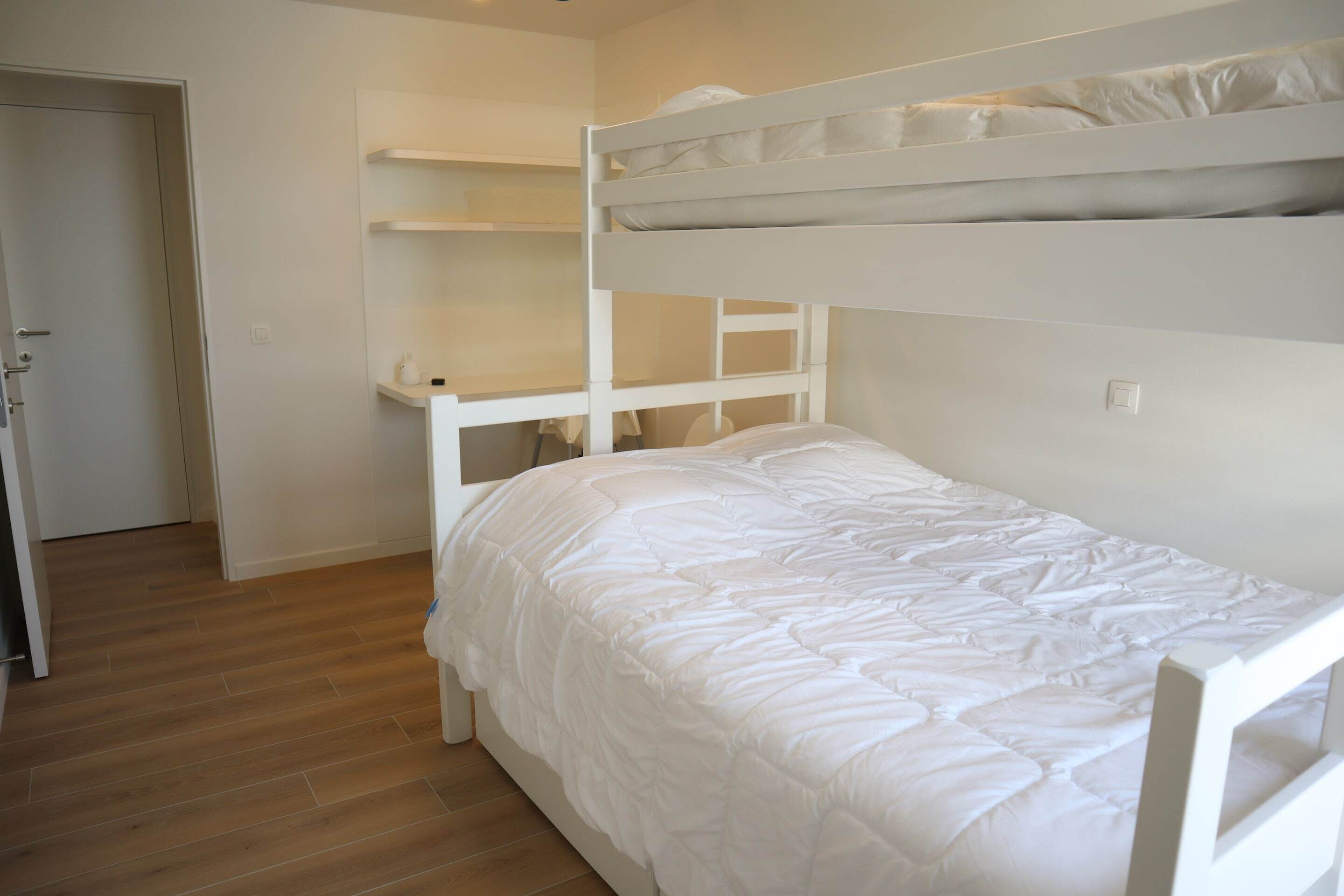 A bunk bed with white bedding, a wooden floor, and white walls.