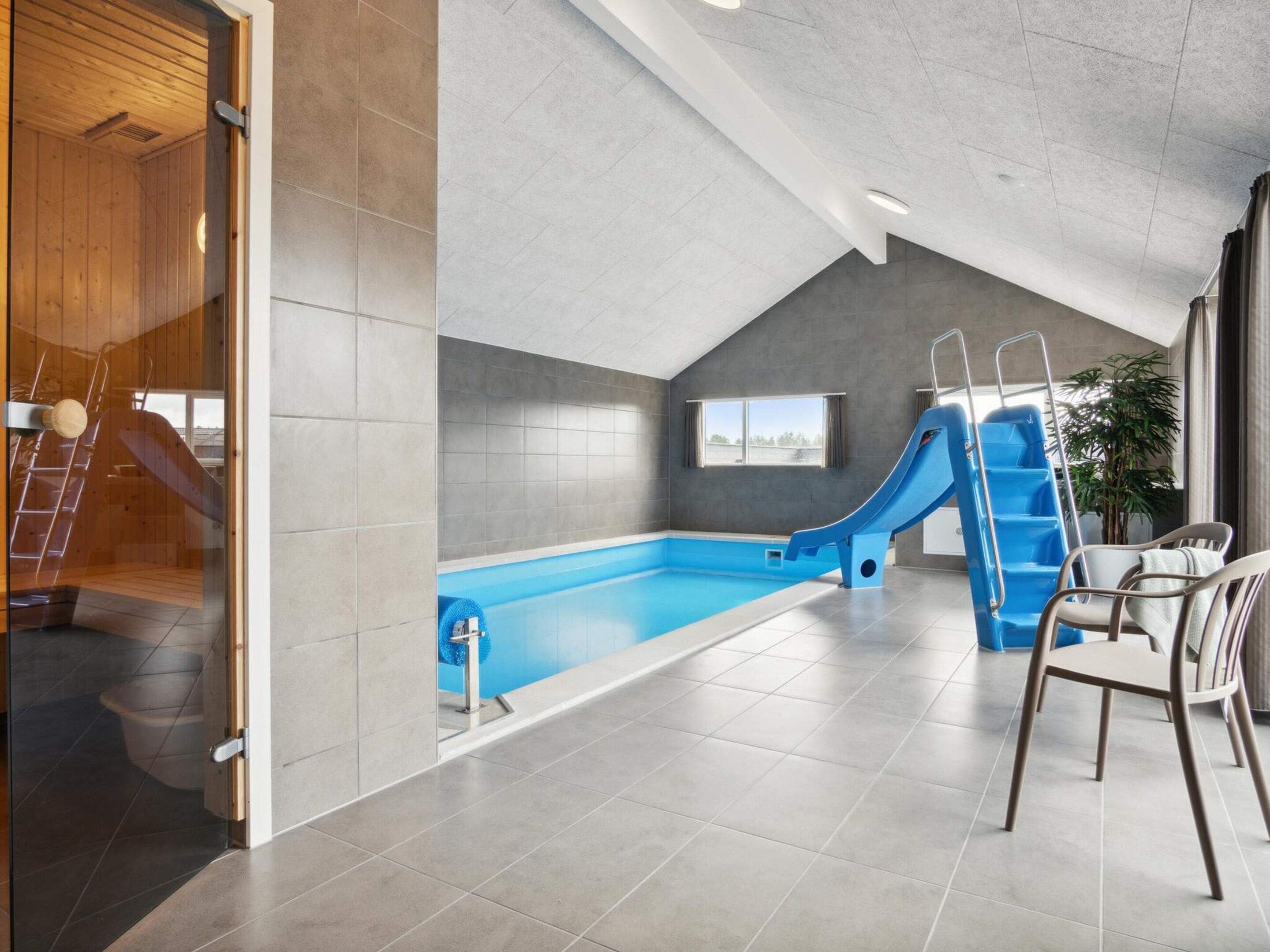 Una piscina interior moderna con un tobogán, una escalera y una zona de estar con sillas.