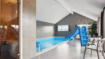 A modern indoor swimming pool with a slide, a ladder, and a seating area with chairs.