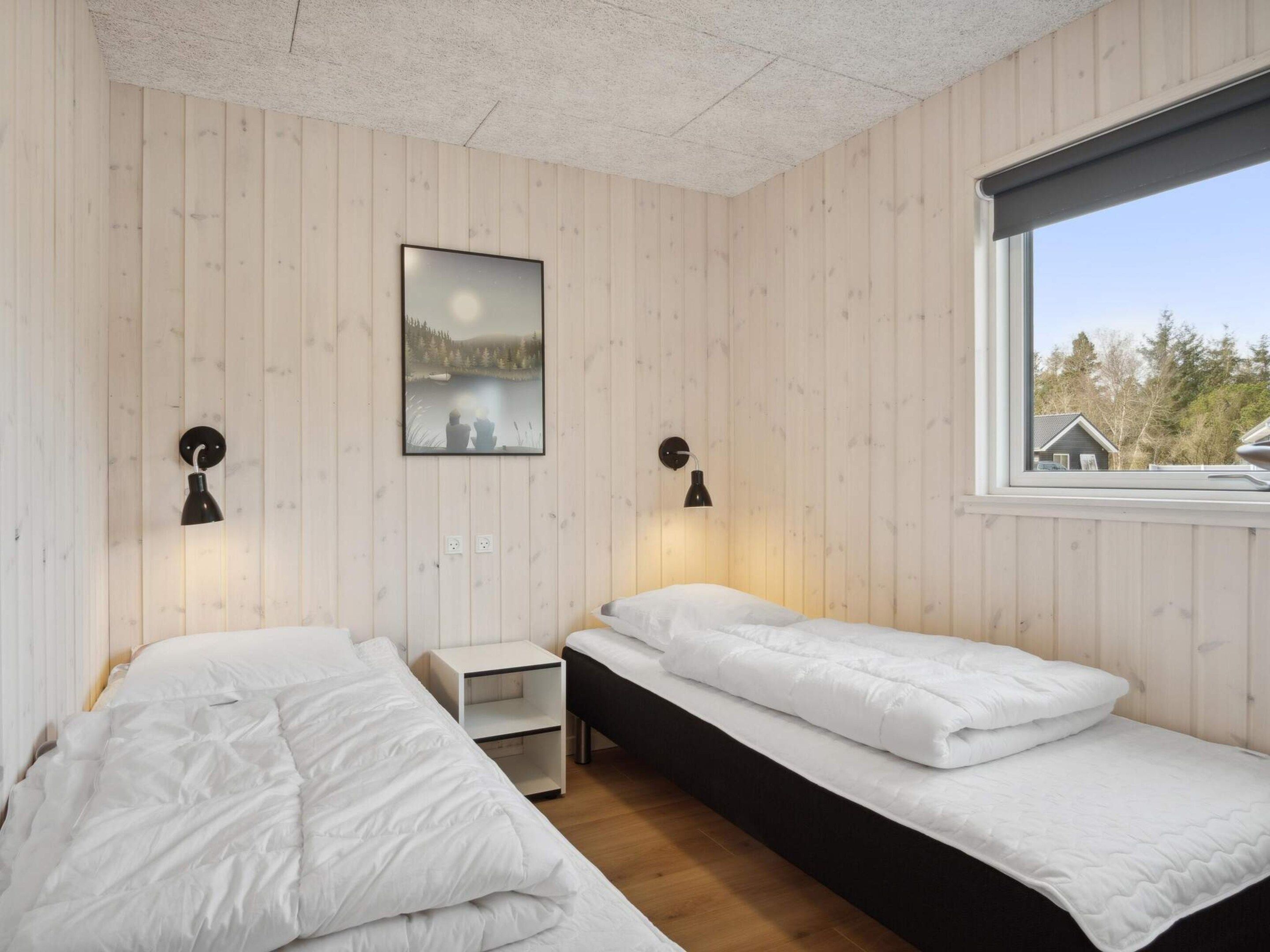 A room with two beds, a nightstand, a wall-mounted picture, and a window with a view of trees.