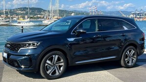 Un SUV noir est garé au bord de l’eau, avec des bateaux et des grues en arrière-plan.