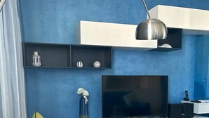 Un salon moderne avec un mur bleu, un téléviseur à écran plat, une étagère noire ornée d’objets décoratifs et un luminaire suspendu.