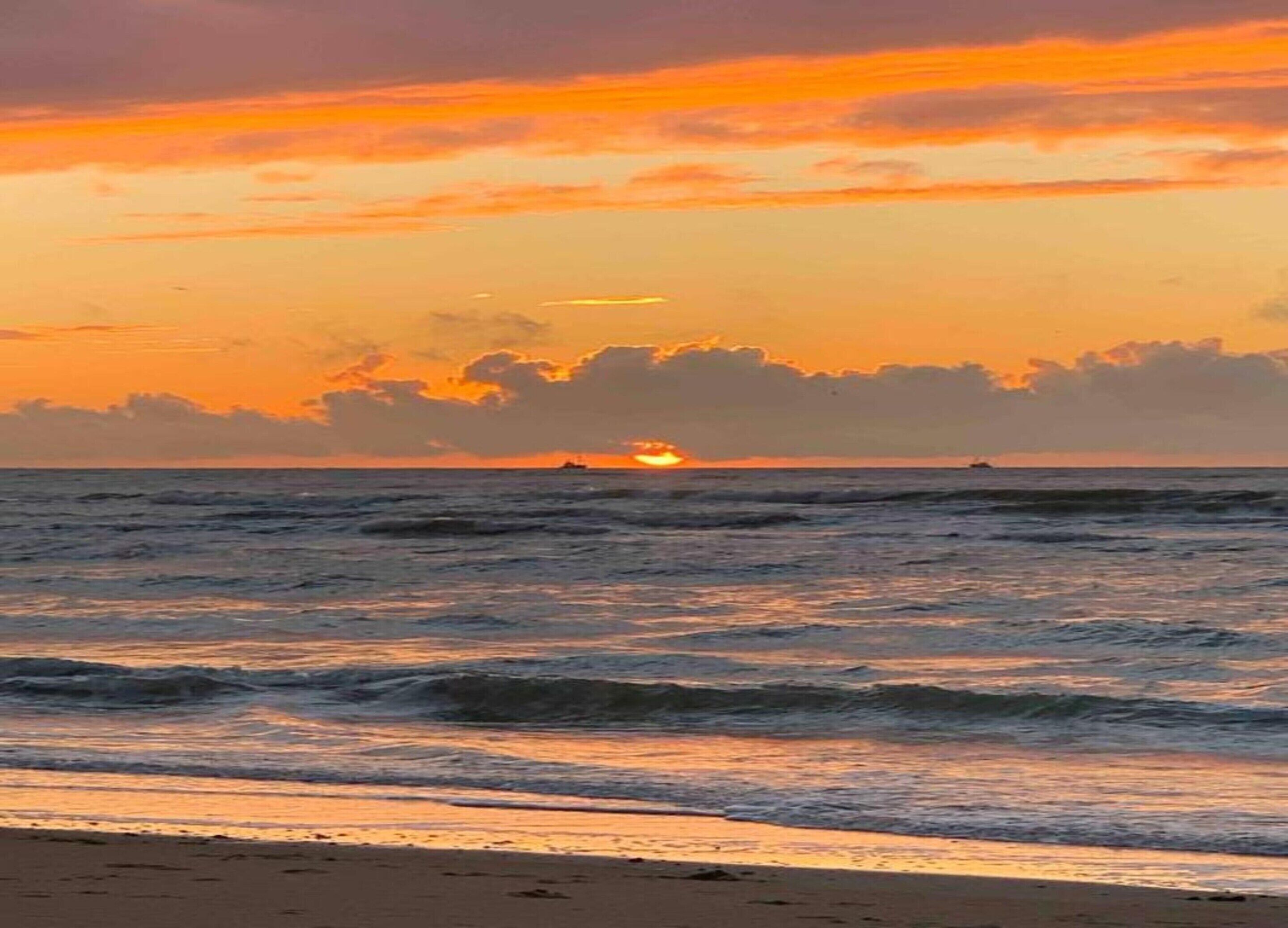 Ein Strand bei Sonnenuntergang mit Wellen und einem Schiff in der Ferne.