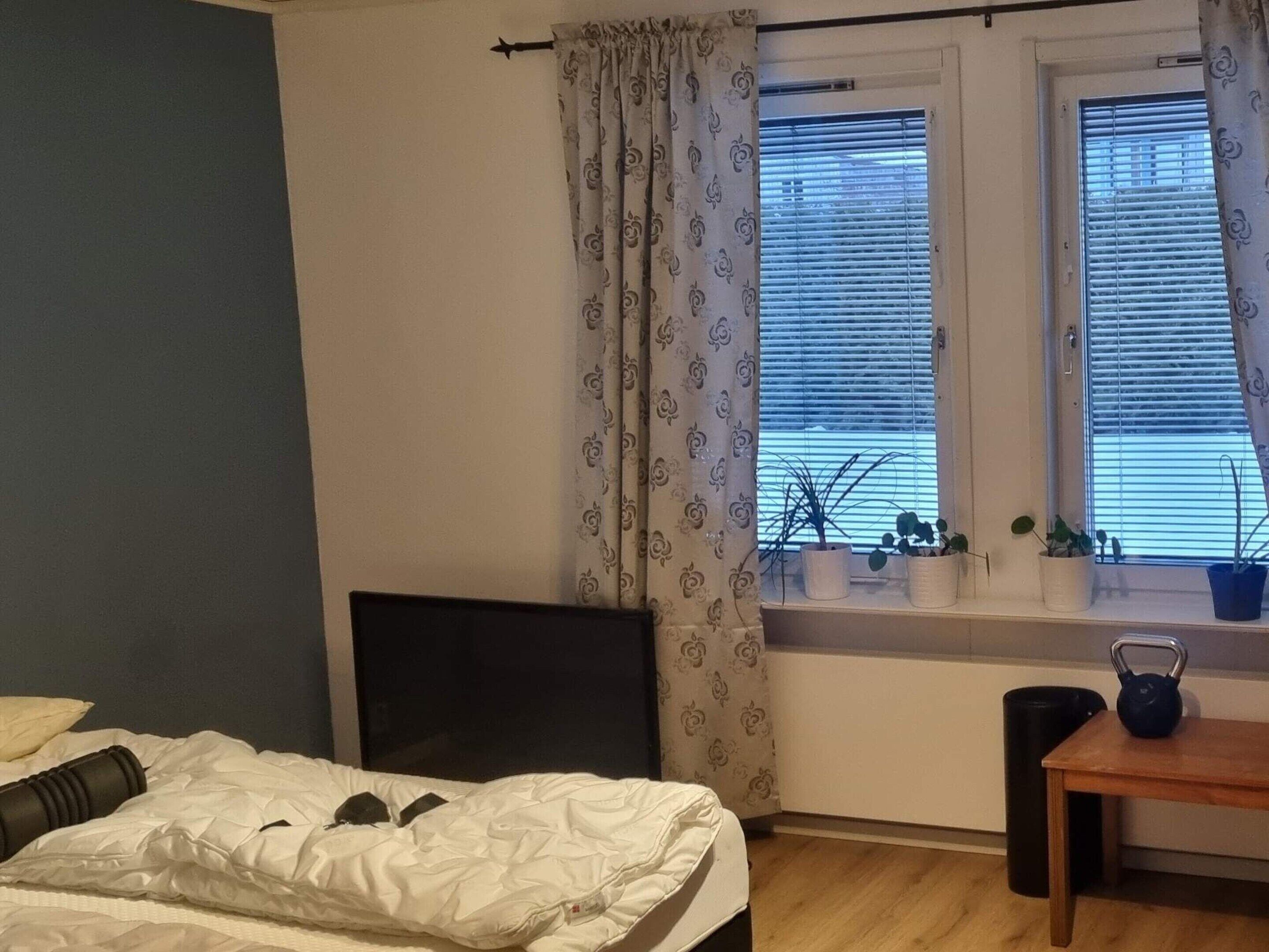 Un dormitorio con cama, ventana con persianas, una mesita con hervidor y un radiador.