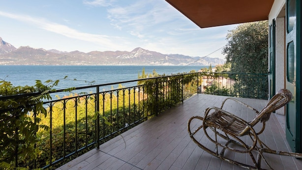 Un balcon avec un fauteuil en osier, une vue sur un lac et des montagnes en arrière-plan.