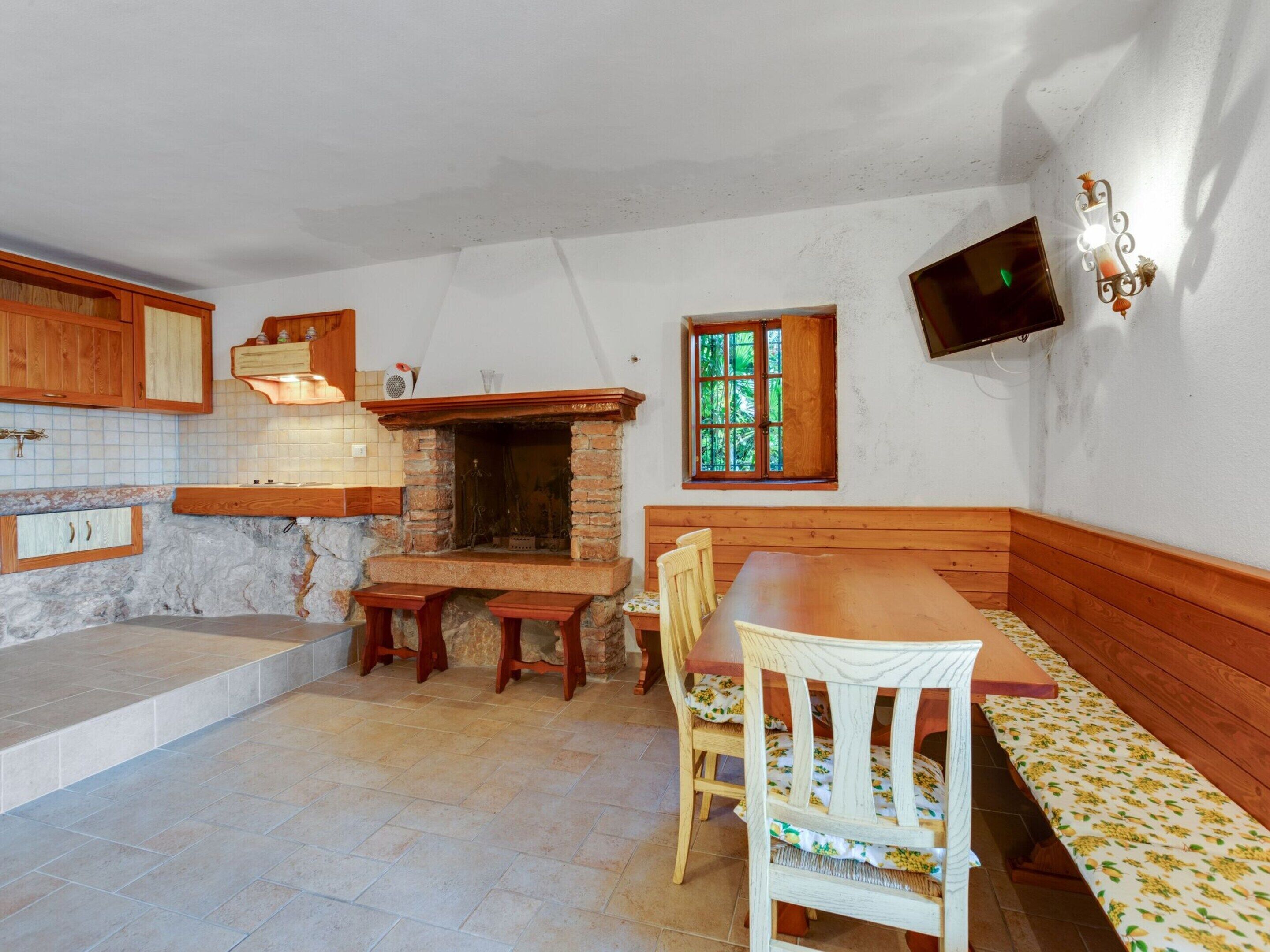 A kitchen with a stone wall, wooden cabinets, a built-in oven, a dining table with benches, and a window with green shutters.