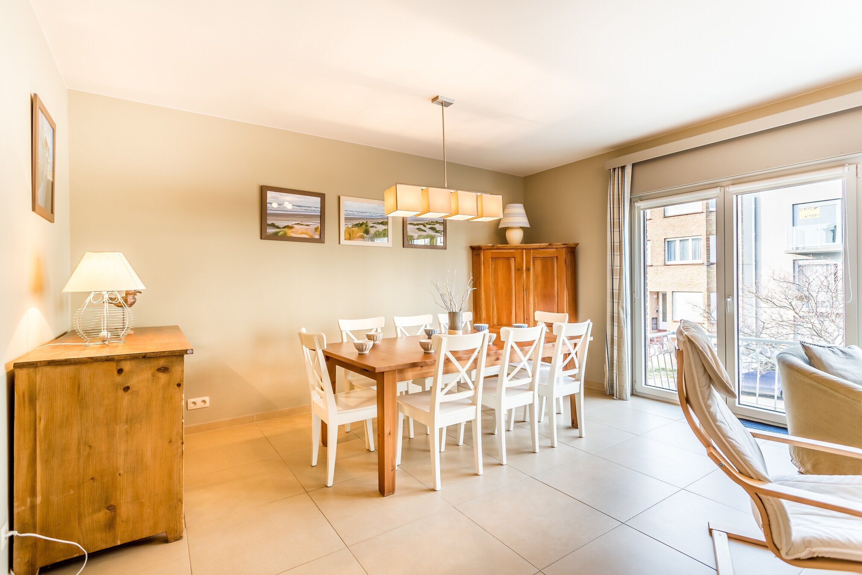 A dining area with a wooden table and white chairs, a wooden cabinet, and a lamp.