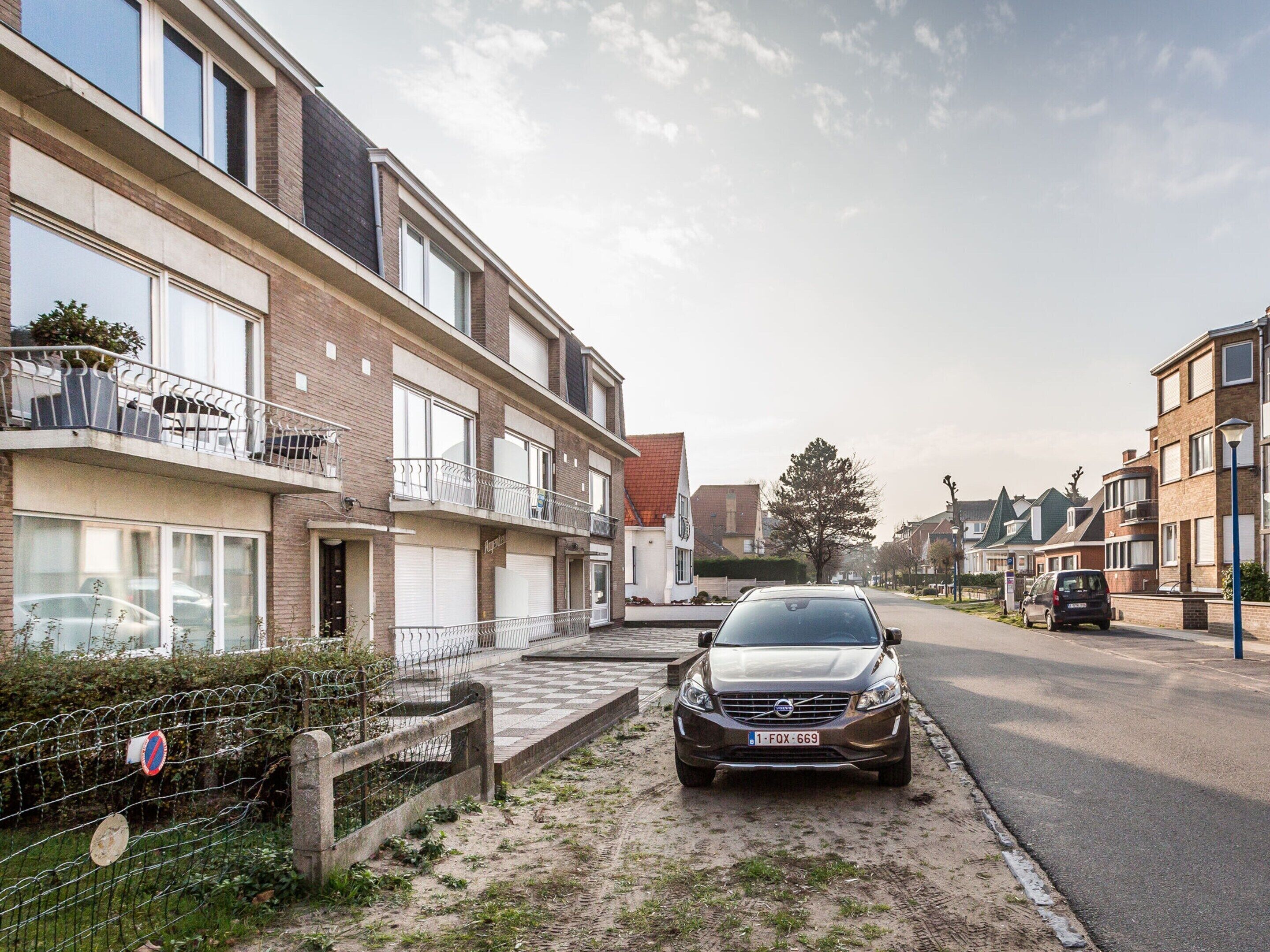 Een woonstraat met een geparkeerde auto, huizen met balkons en een heldere lucht.