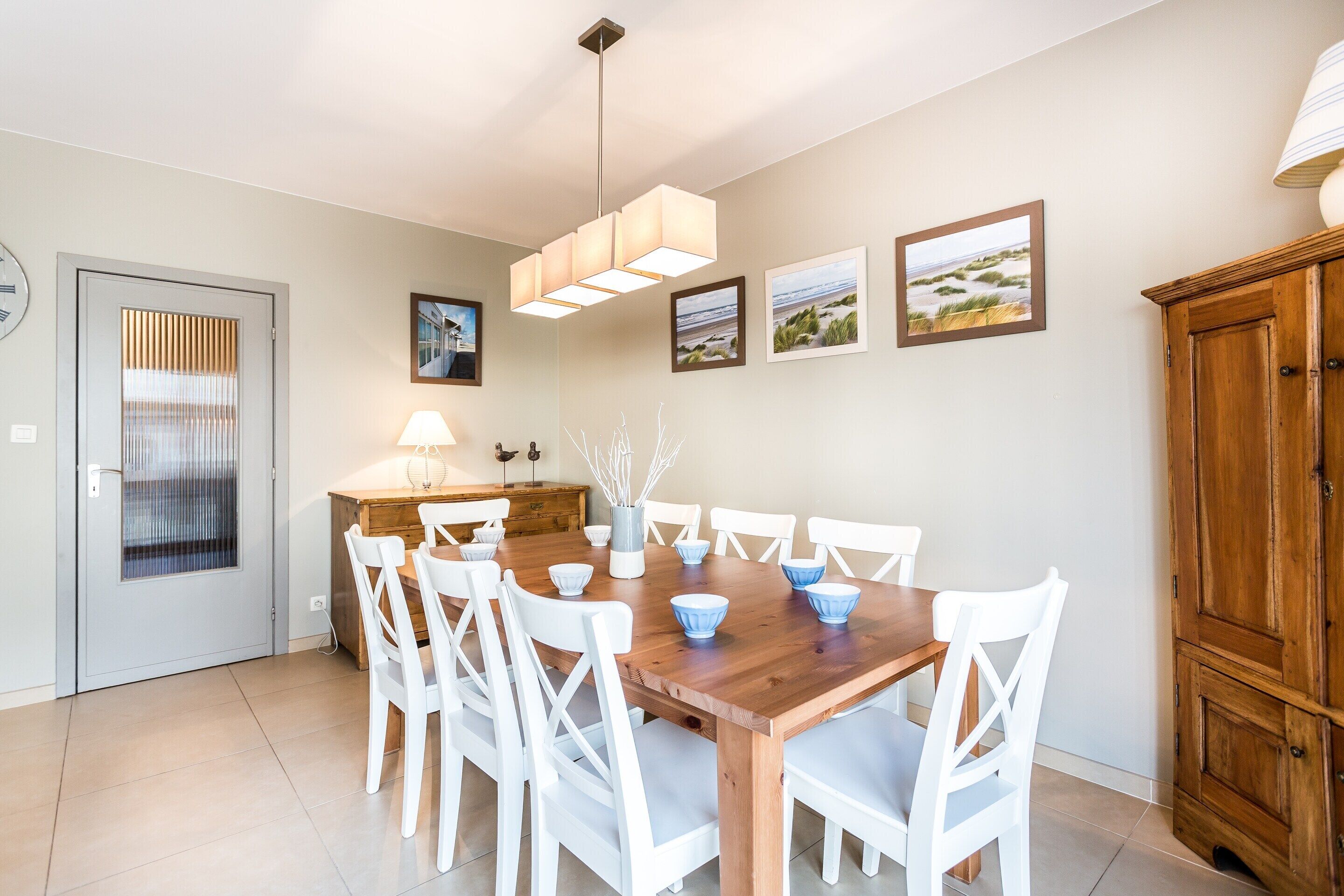 Uma área de refeições com mesa de madeira e cadeiras brancas, luminária pendente e quadros nas paredes.