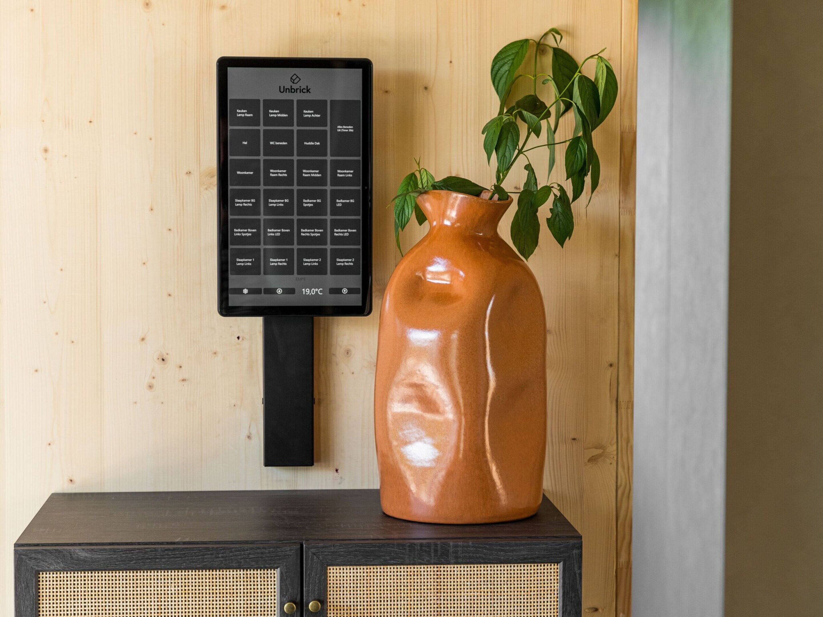 A Unbrick thermostat on a wooden wall, a ceramic vase with a plant, and a dark wooden cabinet with a woven front panel.
