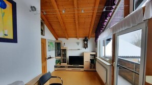 Een gezellige woonkamer met een houten plafond, een televisie, een boekenkast en een groot raam.