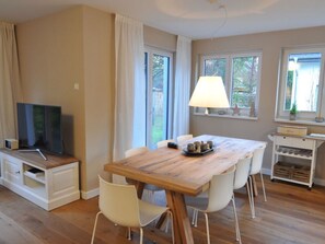 A dining area with a wooden table, white chairs, a television on a cabinet, and a lamp on a side table. - Holiday House Dune Forest b (Ostseebad Dierhagen)