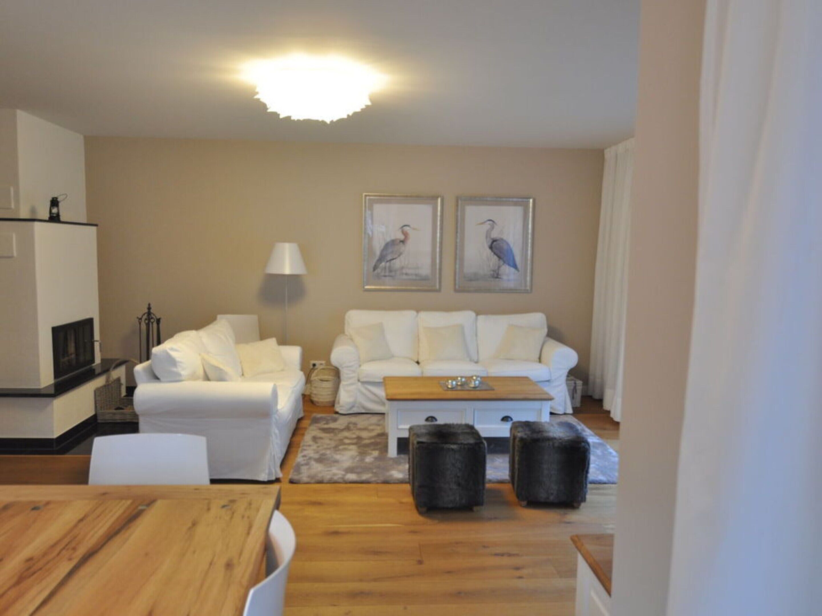 Uma sala de estar moderna com um sofá branco, uma mesa de centro de madeira e dois pufes. Há uma lareira, um abajur e duas molduras na parede.