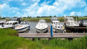 Une rangée de bateaux est amarrée à un quai, avec un pré verdoyant au premier plan et un ciel dégagé.