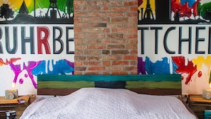 A bedroom with a brick wall, a bed, and a nightstand with a lamp and a book. - Cosy Apartment in Duisburg (Duisburg)