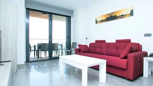 A modern living room with a red sofa, a white coffee table, and a glass door leading to a balcony with a table and chairs. - Belvilla by OYO Apartamento 2habitaciones (Fuengirola)