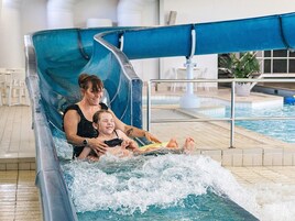 A water slide with a person and a child at the bottom, indoor pool, and seating area.