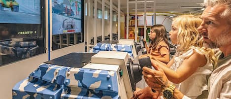 Una habitación con una pantalla grande mostrando un videojuego, asientos con cajas de estampado camuflado y una persona sosteniendo un control de juego.