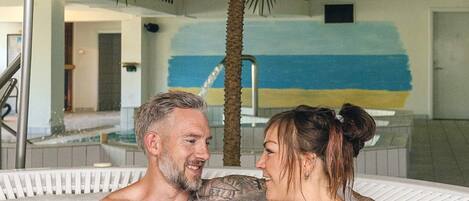 A couple in a hot tub with a palm tree and a blue and yellow wall in the background.