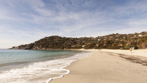 Une plage avec un rivage sablonneux, une eau calme et une colline en arrière-plan, parsemée de bâtiments.