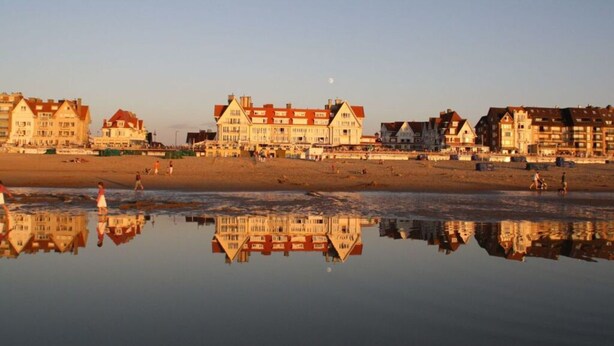 A beachfront hotel with multiple buildings, a reflective water surface, and a clear sky. - Schmitz Comfortable Holiday Residence (De Haan)