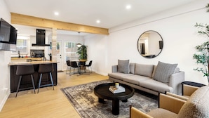 A modern living room with a sofa, coffee table, and chairs. There is a kitchen area with a dining table and chairs. A large mirror hangs on the wall. - Cute 3BR/2BA w/ Jacuzzi, Steps from Big House! (Ann Arbor)