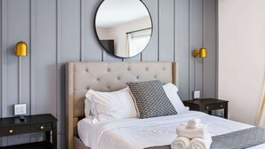 A neatly arranged hotel room with a tufted headboard, a round mirror, two bedside tables with lamps, and a neatly made bed with rolled towels. - Cute 3BR/2BA w/ Jacuzzi, Steps from Big House! (Ann Arbor)
