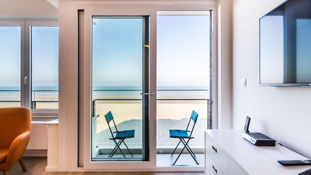 A modern room with a large sliding glass door leading to a balcony with beach views. There is a desk with a computer and a TV mounted on the wall. - Apartment on Zeedijk With Stunning Sea View (Koksijde)
