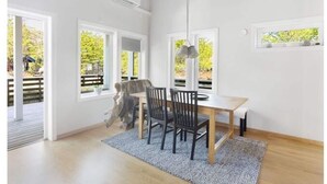 Ett ljust och rymligt rum med ett matbord i trä, två stolar och en hängande lampa. Stora fönster släpper in naturligt ljus och ger utsikt över grönområden utanför.